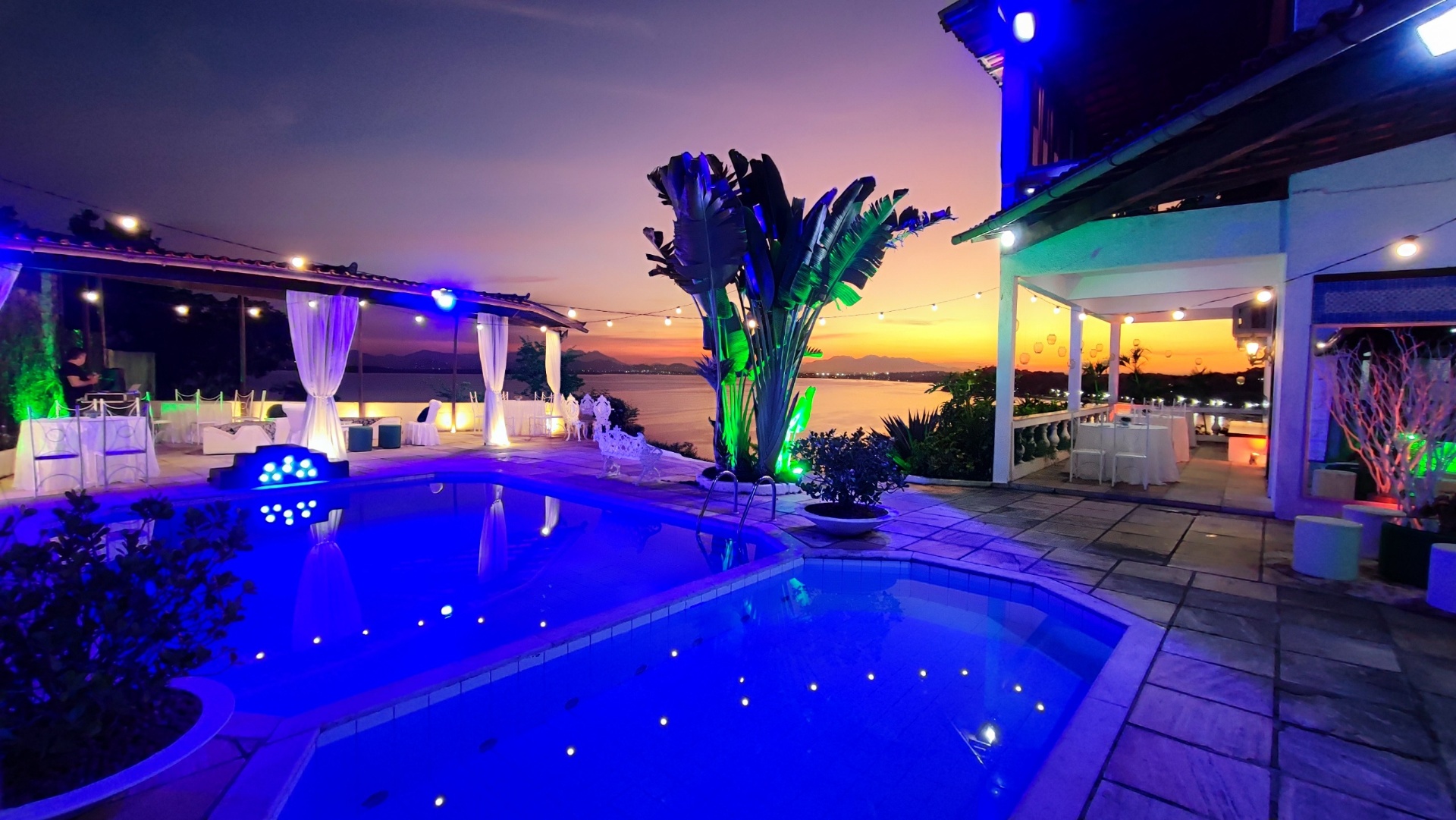Piscina iluminada ao entardecer na Casa Tropical Festas com vista para o mar e decoração de eventos